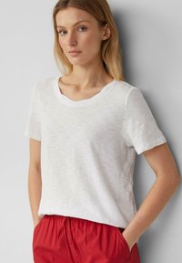 T-shirt blanc à manches courtes avec une finition légèrement texturée et un décolleté en V, associé à un short léger rouge, les mains dans les poches.