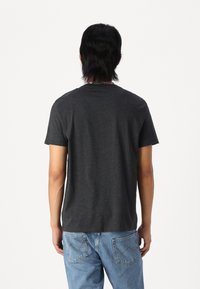 Personne aux cheveux noirs mi-longs portant un t-shirt gris foncé à manches courtes et un jean bleu clair, de dos sur un fond blanc.