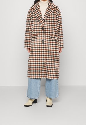 Sprawdzony płaszcz w kratę w brązowych, kremowych i rdzawych kolorach, z kołnierzem o wyprofilowanych krawędziach, dwoma guzikami oraz bocznymi kieszeniami. Noszony na szerokich jeansach.