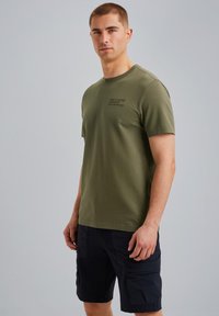 Camiseta de algodón color verde oliva con mangas cortas, que presenta un cuello redondo y un pequeño logo impreso en el lado izquierdo del pecho. Llevado con pantalones cortos cargo de color azul marino oscuro.