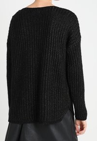 Svart stickad tröja med en texturerad, ribbad design. Har en avslappnad passform, bred ringning och längre bakre fåll.