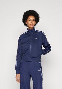 Tuta da ginnastica blu navy con giacca con zip e polsini elasticizzati, caratterizzata da strisce laterali bianche e un logo verde. Texture liscia, design aderente.