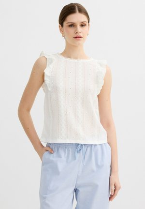 Joven mujer que lleva una blusa blanca sin mangas con volantes y pantalones azul claro con cordón, de pie con una mano en el bolsillo.