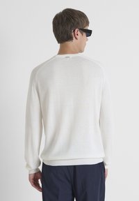 Suéter de punto blanco con mangas largas y detalles acanalados en los hombros. Textura suave, corte informal y dobladillo ligeramente curvado en la parte trasera.