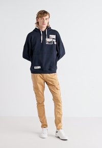 Mörkblå sweatshirt med vita dragband och etiketter, matchad med bruna cargobyxor. Modellen bär vita sneakers och står framför en enfärgad bakgrund.