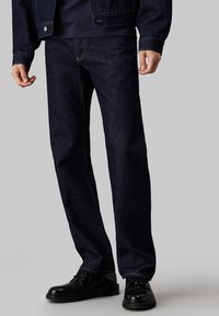 Mörkblå denimjeans med raka ben, synliga sömmar och en klassisk femficksdesign, kombinerade med svarta skor.