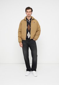 Καστανό bomber jacket με φερμουάρ μπροστά, σε συνδυασμό με ναυτικό μπλε πουλόβερ με ρόμβους και μαύρο τζιν. Λευκά αθλητικά παπούτσια ολοκληρώνουν την εμφάνιση.