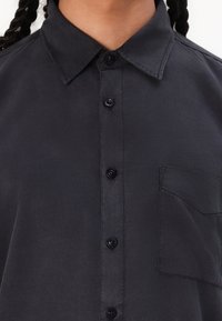 Marineblaues Hemd mit Knöpfen aus glattem Stoff, das einen klassischen Kragen, sechs Vorderknöpfe und eine einzelne Brusttasche aufweist.
