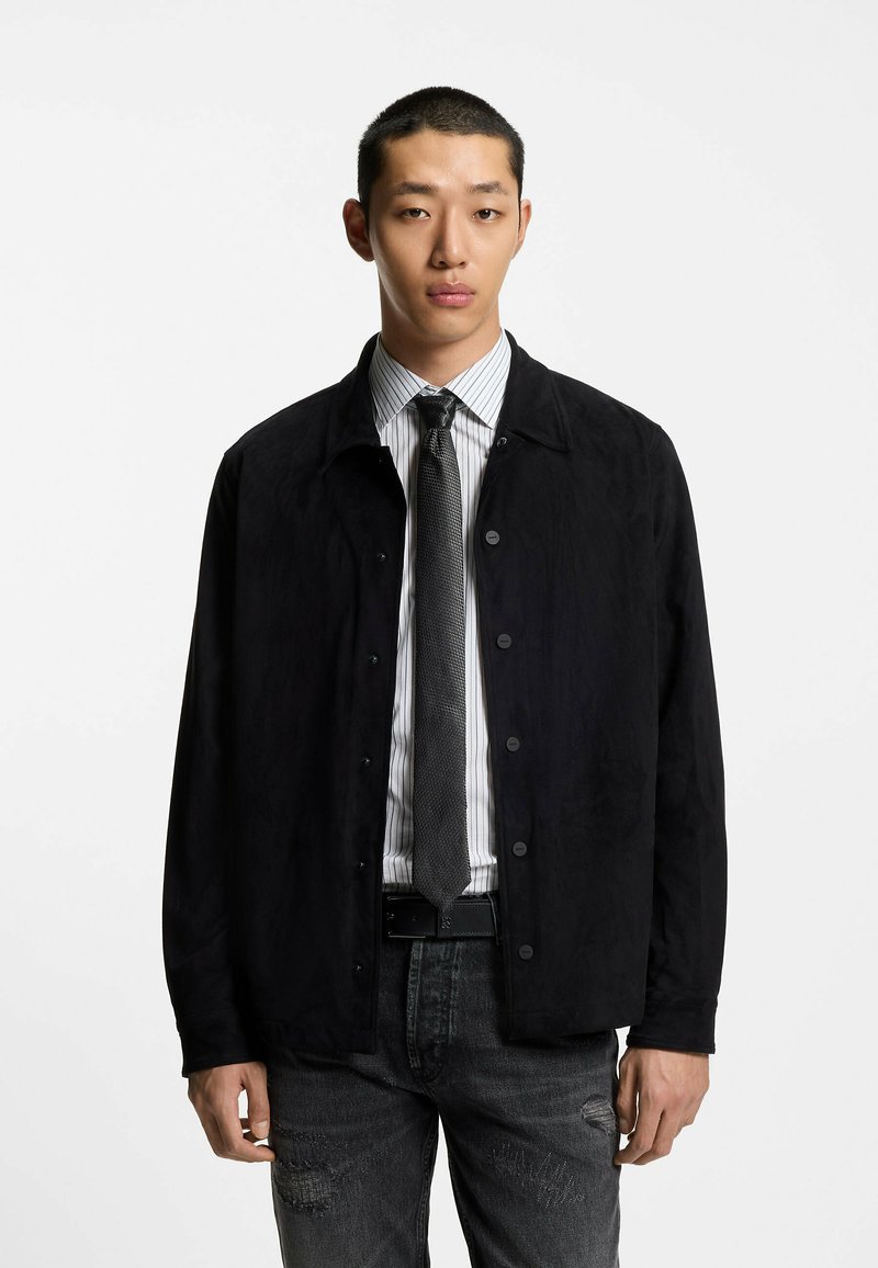 Giacca in suede nera con collo a risvolto, davanti con bottoni, abbinata a una camicia a righe e cravatta texturizzata. Indossata con jeans scuri usurati.