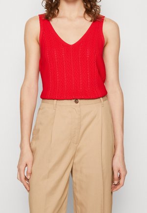 Czerwony top bez rękawów z dzianiny z dekoltem w serek, zestawiony z beżowymi spodniami z wysokim stanem i plisami, prezentowany na stojącej osobie, od ramion do bioder.