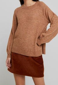 Pull en maille marron à col rond, coupe décontractée et poignets côtelés, associé à une mini-jupe en velours côtelé rouille avec des côtes verticales.