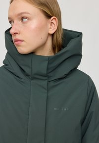 Grüne gepolsterte Jacke mit hohem Kragen, nahtlosem Design und dezentem Logodetail. Das Material wirkt glatt und wasserabweisend.