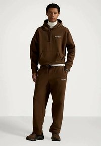 Νεαρός άνδρας φορώντας καφέ φούτερ Polo Sport και αντίστοιχες φόρμες, στέκεται με το ένα χέρι στην τσέπη μπροστά σε μονόχρωμο φόντο.