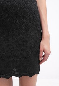 Černé krajkové šaty s texturovaným květinovým vzorem a vlnkovým lemem. Detail dolní části a ruka spočívající na látce.