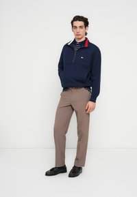 Μπλε πουλόβερ με λεπτομέρειες κόκκινου στο γιακά, σχεδίαση με μισό φερμουάρ και μπαλώμα με λογότυπο. Μπεζ παντελόνια και γυαλιστερά μαύρα παπούτσια ολοκληρώνουν το σύνολο.