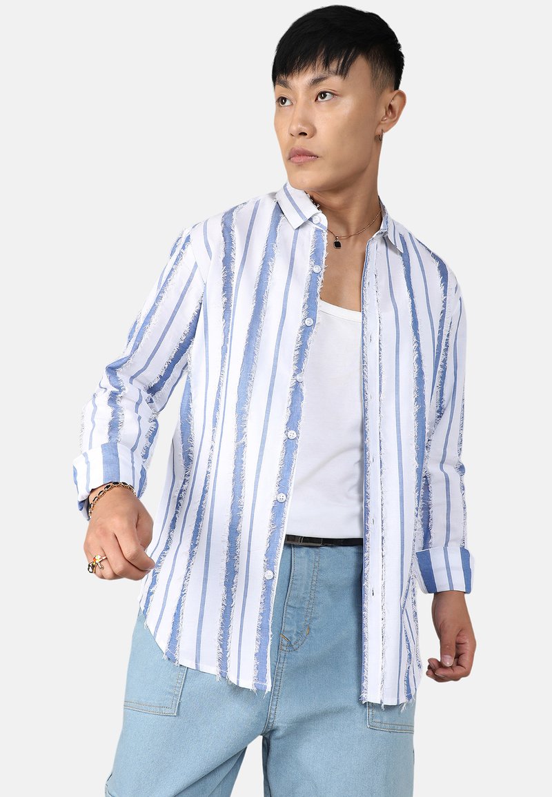 Blau-weißt gestreifte Bluse mit geknöpfter Vorderseite, weichem Material, fransigen Kanten, langen Ärmeln und lässigem Schnitt. Getragen über einem weißen Top.