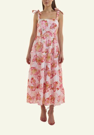 Frau trägt ein ärmelloses, weißes Maxikleid mit pinkfarbenem Blumenmuster und gebundenen Trägern sowie pinkfarbene Riemchenabsätze.