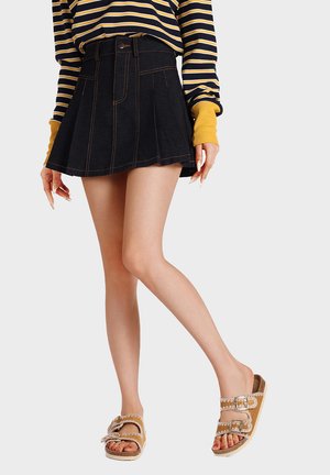 Donna con gonna nera plissettata corta, top a maniche lunghe a righe con polsini gialli e sandali color cuoio con doppia fibbia.