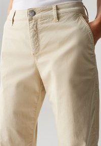Beige Baumwoll-Chinos mit glatter Textur, ausgestattet mit einem Reißverschluss und Knopfverschluss vorne, Gürtelschlaufen und Seitentaschen. Sichtbare Steppnaht.