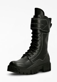 Bota de combate de cuero negro con detalles de tachuelas, cordones delanteros y suela gruesa. Presenta una correa decorativa alrededor del tobillo.