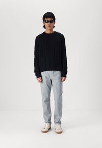 Marineblauer Kabelstrickpullover mit langen Ärmeln, kombiniert mit hellgrauen Hosen und weißen Sneaker mit braunen Sohlen. Dunkle Sonnenbrille getragen.