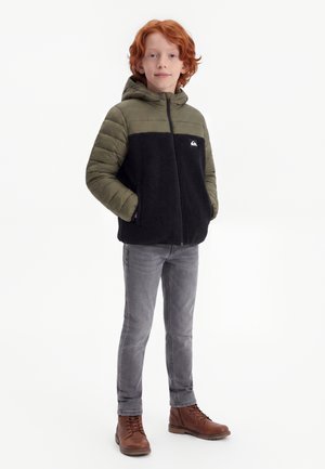 Enfant aux cheveux roux portant une veste à capuche olive et noire, un jean gris et des bottines marron à lacets, debout les mains dans les poches.