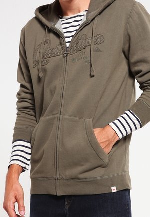 Person iført olivengrøn hættetrøje med lynlås og Quiksilver-logo, lagdelt over en sort- og hvidstribet langærmet skjorte, hånd i lommen.