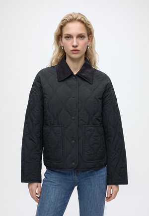 Kvinde med blondt hår iført en sort quiltet jakke med en mørk krave og blå jeans, stående mod en ensfarvet lys baggrund.