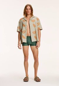 Lühikeste varrukatega helesinine särk oranžide lehemustritega, tumeroheliste lühikeste pükstega ja pruunide sandaalidega. Modell seisab sirgelt.