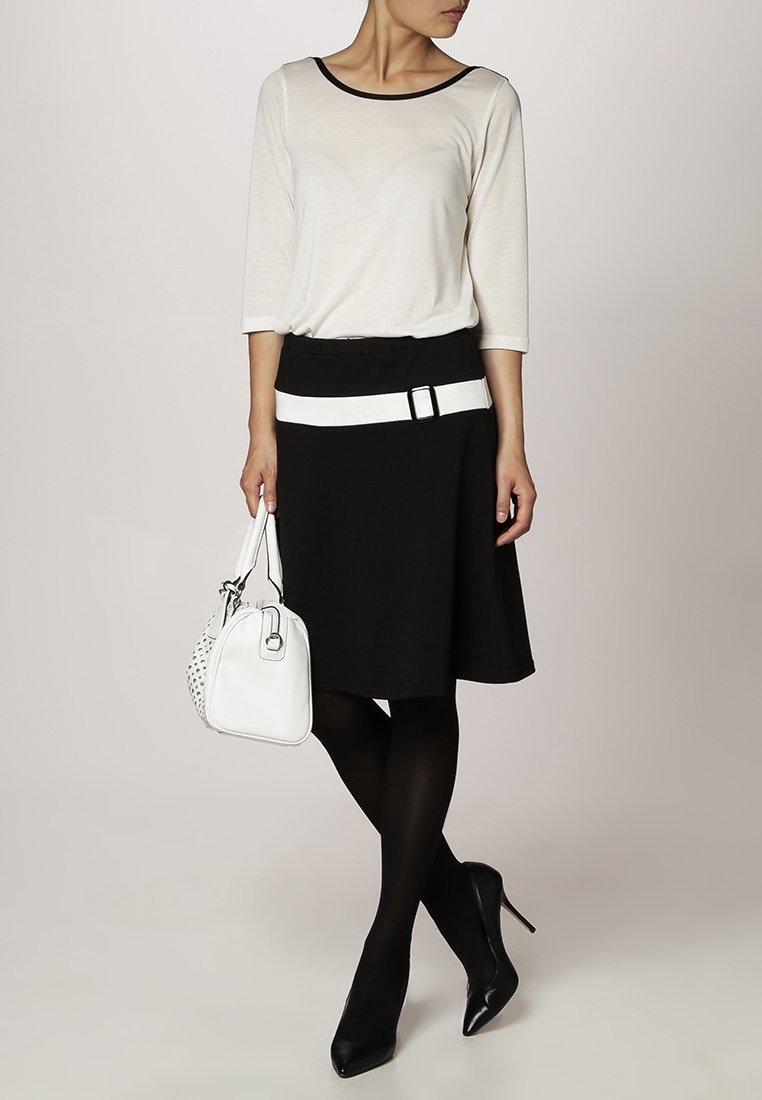 Femme portant un haut blanc avec bordure noire, jupe noire avec ceinture blanche, collants noirs, talons noirs, tenant un sac à main blanc.