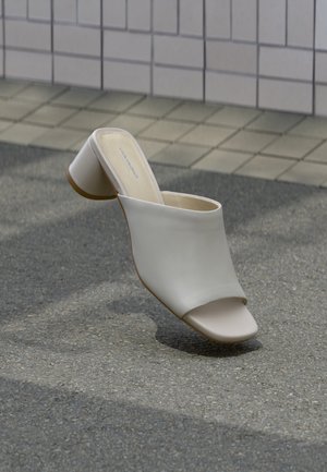 Bijela ženska natikača otvorenih prstiju s blok petom na popločanoj površini uz zid od pločica, položena tako da se vide bočni i gornji prikazi.