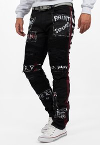 I pantaloni in denim neri presentano dettagli strappati, spiccate grafiche in bianco e rosso, sezioni di tessuto a coste testurizzate e rifiniture in metallo argentato.