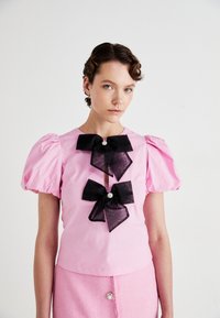 Kvinne med kort krøllete hår iført en rosa bluse med pufferm og to store svarte sløyfer, samt et rosa skjørt med en dekorativ knapp.