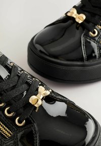 Schwarze Lackledersneaker mit goldenem Hardware-Zubehör, strukturierten Schnürsenkeln und einer dekorativen Schleife. Verfügt über eine glänzende Oberfläche und eine Gummisohle.