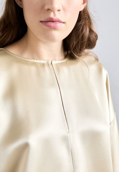Satin blouse in lichtbeige met een ronde halslijn en een discrete sluiting aan de voorkant. Glad textuur met subtiele glans en minimale stikseldetails.