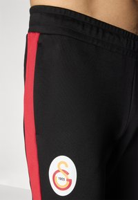 Sorte træningsbukser med en rød sideliste, der har et cirkulært logo med "S" og "1905" i orange og hvid på nederste del af benet.
