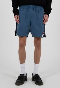 Mann trägt blaue Shorts mit Seitenstreifen, weiße Kniestrümpfe, schwarze Loafers und ein schwarzes Langarmshirt vor einem schlichten Hintergrund.