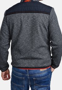 Muž v tmavě šedém strukturovaném saku s černým ramenním panelem a modrými džíny, zobrazený zezadu s jednou rukou v kapse.