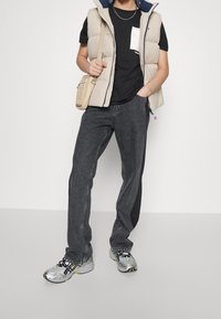 Beige vadderad väst över en svart t-shirt, grå jeans och silverfärgade sportskor med svarta detaljer. Liten ljus väska bärs över kroppen.