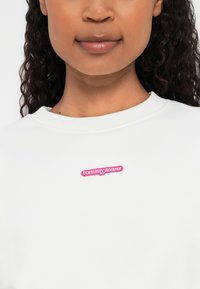 Valge sviiter ümmarguse kaelusega, mille ees on roosa "tommy jeans" logotempel. Pehme kangas sujuva tekstuuriga.