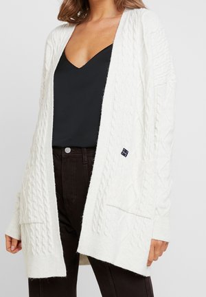 Femme portant un cardigan blanc ouvert en maille torsadée sur un top noir à col en V et un pantalon foncé, debout devant un fond uni.