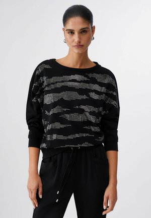 Donna con capelli tirati indietro che indossa una felpa nera con motivo mimetico con borchie argentate e pantaloni neri con coulisse.