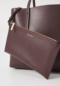 Sac fourre-tout en simili cuir bordeaux avec deux poignées, doté d'une pochette zippée amovible avec quincaillerie dorée et logo "Calvin Klein" en relief.
