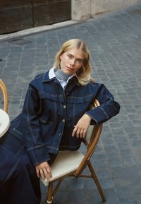 Dunkle Jeansjacke mit heller Naht, Knopfverschluss und Brusttaschen, kombiniert mit einem grauen Rollkragenpullover und einem weißen Hemd, sitzt auf einem Stuhl.