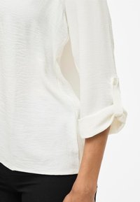 Hvid, tekstureret bluse med en afslappet pasform og opsmøgede ærmer, der har en knapdetalje på ærmerne. Båret over sorte bukser.