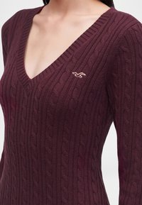 Femme portant un pull en maille torsadée bordeaux foncé avec un col en V et un petit logo brodé de mouette sur la poitrine.