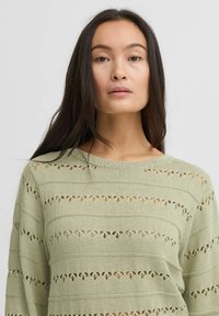 Stickad tröja i ljusgrön med en strukturerad design med horisontella ränder och utskärningar. Rund halsringning och långa ärmar.