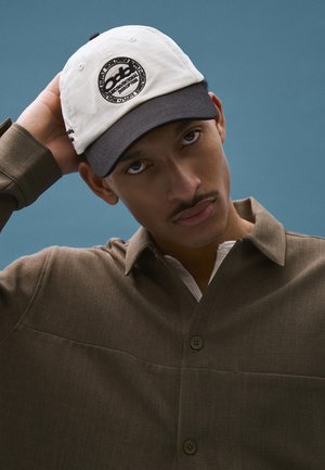 Mladý muž s knírem, který nosí bílou a černou čepici a hnědou košili na knoflíky, dotýká se zadní části hlavy na modrém pozadí.