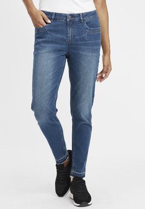 Blaue Jeans aus Denim mit Slim-Fit, ausgestattet mit Fronttaschen und einem ausgefransten Saum. Kombiniert mit schwarzen Sneakers für einen lässigen Look.