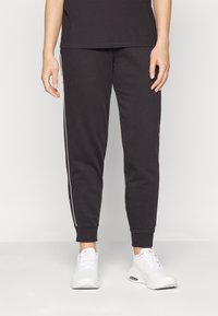 Calças de moletão pretas com riscas brancas nas laterais, punhos ajustados e um acabamento de tecido suave. Estilizadas com sapatilhas atléticas brancas.